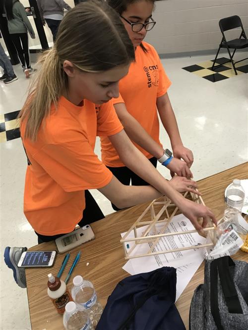 Two girls wearing orange STEM shirts standing at a table building a bridge from wood sticks. 